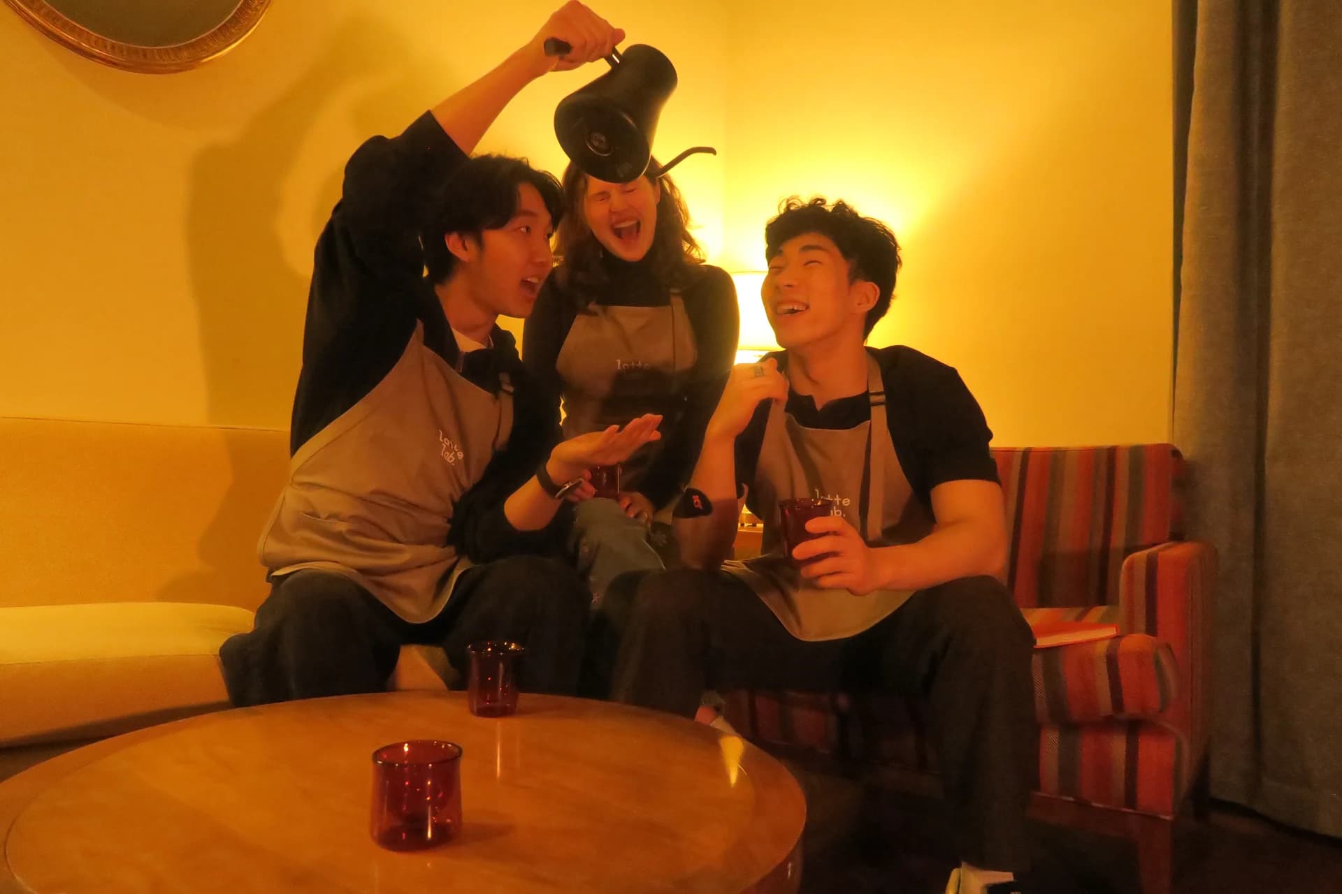 Three Latte Lab friends in aprons laughing together on a couch, one holding a kettle
